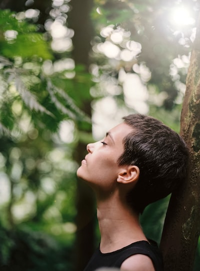 "How to Build Resilience for Better Mental Health" Women with short hair, leaning against a tree in the woods, looking tired bur relieved